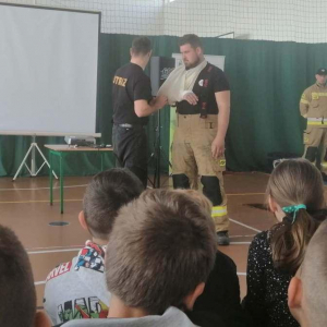 Strażacy prezentują jak założyć chustę by unieruchomić rękę w przypadku złamania lub zwichnięcia.