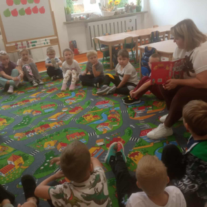 Dzieci siedzą na dywanie i słuchają jak czyta Pani Bibliotekarka.